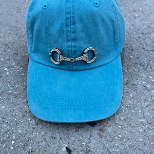 May include: A light blue baseball cap with a silver and rhinestone equestrian bit design.