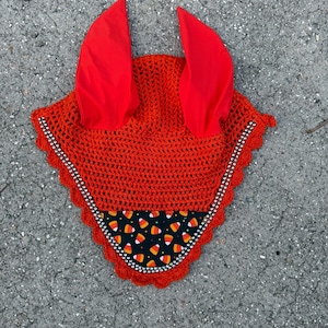 May include: A bright orange crochet horse bonnet with two red fabric horns. The bonnet has a black and white candy corn pattern on the front. The edges are trimmed with a decorative border.