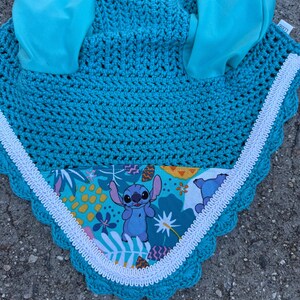 May include: A turquoise crochet horse bonnet with matching ear coverings. The bonnet features a white scalloped trim and a patterned fabric insert with a cartoon character. The ear coverings are made of a smooth, turquoise fabric.