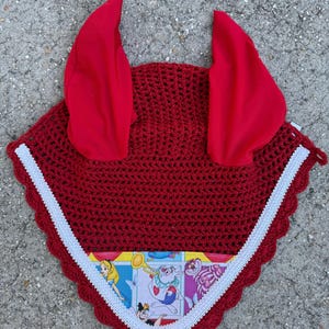May include: A red crochet horse bonnet with red ear covers and a white trim. The bonnet features a colorful fabric panel with illustrations of characters from Alice in Wonderland. This equestrian accessory is designed to protect a horse's ears.