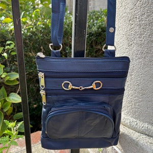 May include: A dark blue leather crossbody bag with multiple zippered compartments and gold-toned hardware. The bag has a long adjustable strap.
