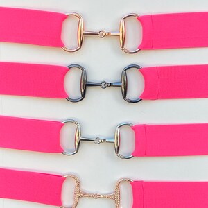 May include: Four pink elastic belts with different colored metal buckles. The buckles are all in the shape of a snaffle bit. The buckles are gold, silver, silver, and gold with rhinestones.