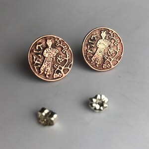 May include: A pair of copper earrings with a detailed design of a person standing in a garden. The earrings are round and have a textured surface. The person in the design is wearing a long robe and holding a fan. The earrings are accented with a gold finish.