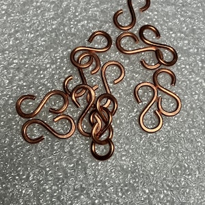 May include: A scattering of small, copper-coloured S-shaped hooks. The hooks are arranged on a textured, light-coloured surface. The hooks are made of metal and have a shiny finish. These hooks are likely used for jewellery making or crafts.
