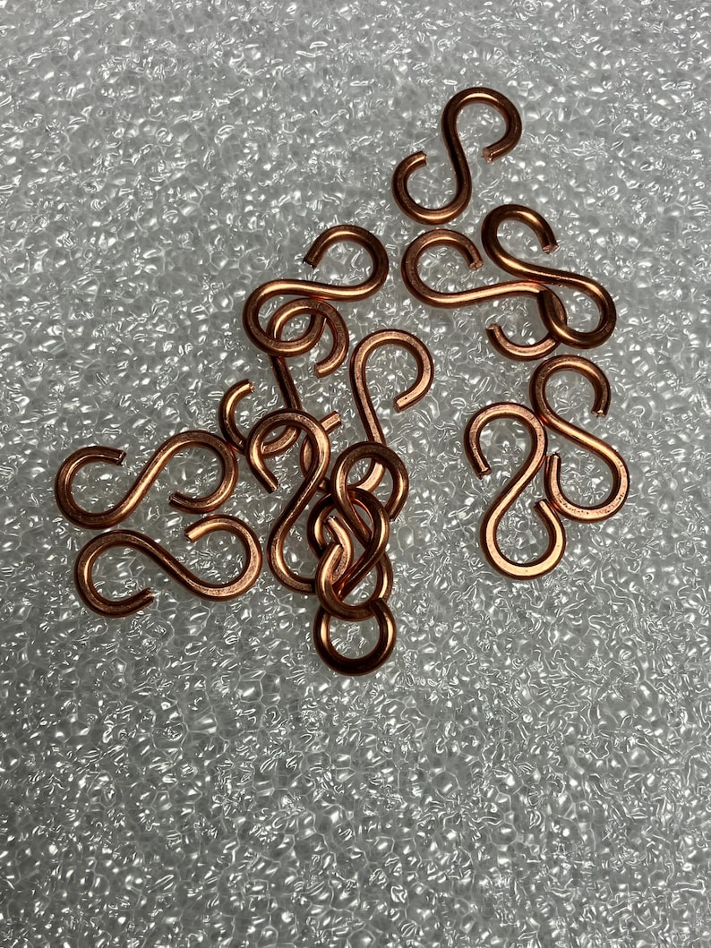 May include: A scattering of small, copper-coloured S-shaped hooks. The hooks are arranged on a textured, light-coloured surface. The hooks are made of metal and have a shiny finish. These hooks are likely used for jewellery making or crafts.