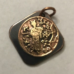 Peut inclure: Un pendentif vintage avec une bélière couleur cuivre. Le pendentif a une bordure en forme de losange gris foncé. Un centre circulaire de couleur or présente un relief détaillé d'une figure et de feuillage.
