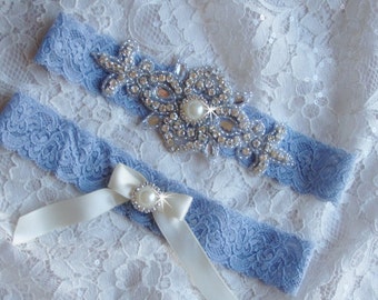 Pode incluir: Um par de ligas de renda azul com um laço de cetim branco e um adorno de strass. O adorno apresenta uma pérola grande no centro.