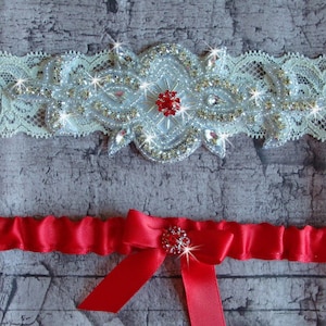 May include: Two bridal garters. The top garter is ivory lace with a central design of silver beads and red and clear rhinestones. The bottom garter is red satin with a bow and rhinestone embellishment.