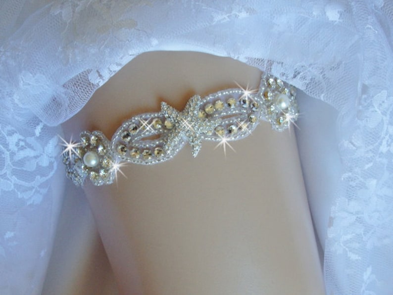 K&ouml;nnte beinhalten: Ein wei&szlig;es Brautstrumpfband mit einem silbernen und kristallenen Design. Das Strumpfband hat eine Perlenverzierung in der Mitte.