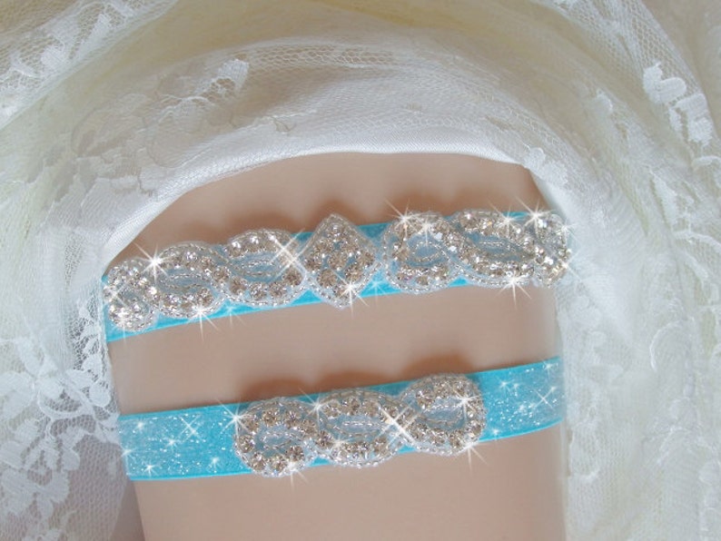 May include: A turquoise blue garter with a silver rhinestone design. The garter is made of satin and has a sparkly finish.