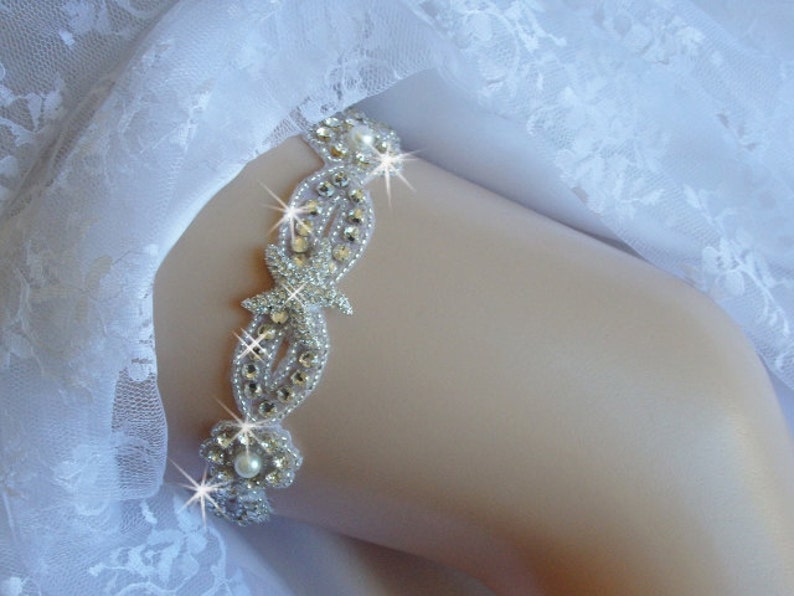 K&ouml;nnte beinhalten: Ein wei&szlig;es Brautstrumpfband mit einem silbernen Strassstein und einer Perle. Das Strumpfband ist mit einem zarten Blumenmuster verziert und hat einen gro&szlig;en, funkelnden Strassstein in der Mitte.
