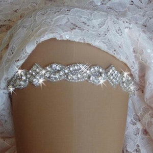 May include: A silver rhinestone garter with a delicate design. The garter is made of a thin band of metal with small, clear rhinestones set in a pattern. The garter is shown on a white lace background.