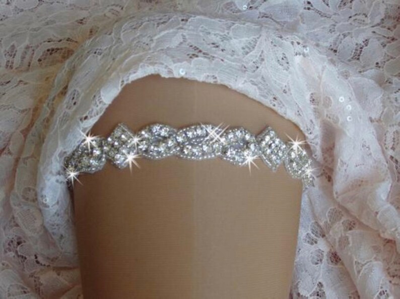 May include: A silver rhinestone garter with a delicate design. The garter is made of a thin band of metal with small, clear rhinestones set in a pattern. The garter is shown on a white lace background.