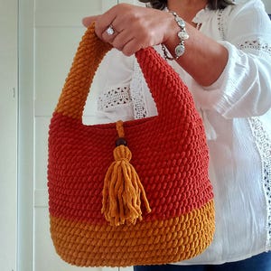 May include: A handmade, two-tone crochet bag in shades of orange and gold. The bag features a textured stitch, a rounded shape, and a matching tassel with a wooden bead accent. The handle is a continuation of the crochet design.