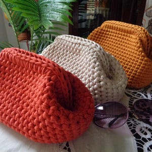 May include: Three handmade crochet pouches in solid colors of burnt orange, cream, and mustard yellow. Each pouch features a thick, textured knit pattern with a structured frame opening. A pair of dark-tinted sunglasses rests near the pouches.