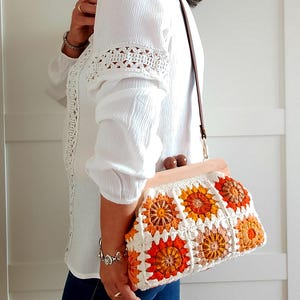 Könnte beinhalten: Cremefarbene Häkeltasche mit Holzrahmen und Griff. Die Tasche hat ein Blumenmuster in Orange- und Gelbtönen. Sie hat einen braunen Lederriemen. Die Tasche ist etwa 30 cm lang.