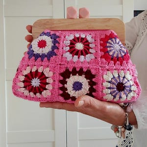 May include: Pink crochet clutch with a wooden frame and pink clasp. The clutch features a floral pattern in red, white, purple, and burgundy. The wooden frame is light brown. A stylish accessory for any occasion.