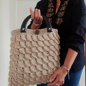 May include: Beige crochet tote bag with dark brown wooden handles. The bag features a textured, scalloped pattern and a wooden button closure. The handles are attached with small, decorative wooden accents. The bag is held by a person wearing a black shirt.
