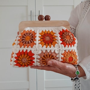 Puede incluir: Un bolso de mano de ganchillo hecho a mano con marco y cierre de madera. El bolso presenta un patrón floral en tonos naranja, melocotón y blanco. El marco de madera tiene dos detalles esféricos marrones. Un accesorio elegante.