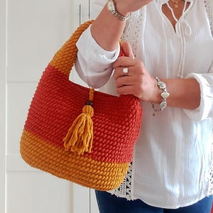 May include: A handmade crochet bag in shades of red and yellow-orange. The bag features a textured stitch pattern, a rounded shape, and a matching tassel. The handle is a continuation of the crochet pattern. A stylish and unique accessory.