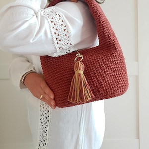 Peut inclure: Un sac à bandoulière crocheté de couleur rouille, de forme arrondie. Le sac est doté d'une bandoulière assortie et d'un pompon décoratif avec des fils dorés et beiges. Un fermoir doré fixe le pompon au sac.