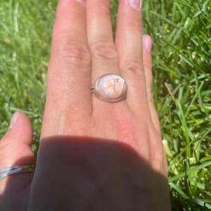 May include: A silver ring with a large oval-shaped stone. The stone is a light pink and white color with brown veins running through it.