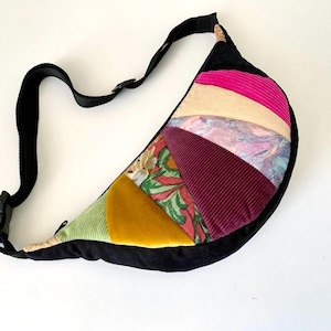 May include: A colourful, patchwork fanny pack with a black adjustable strap. The bag features a variety of fabrics in shades of green, yellow, pink, and burgundy. The bag is approximately 30 cm wide.