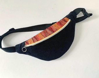 May include: A navy blue corduroy fanny pack with a colorful striped zipper pocket. The zipper is silver, and the adjustable strap is black. The bag is designed to be worn around the waist or across the body.