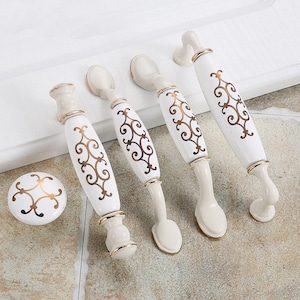 May include: Five white ceramic cabinet knobs with gold accents. The knobs feature a decorative, ornate design.