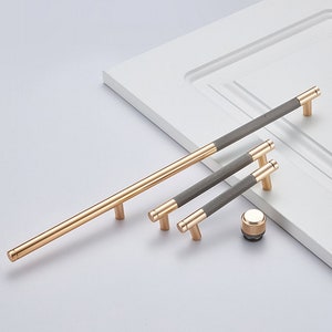 Könnte beinhalten: Ein Satz von vier goldenen Schrankgriffen mit grauer, strukturierter Oberfläche. Die Griffe sind in verschiedenen Größen und auf einer weißen Oberfläche angeordnet.