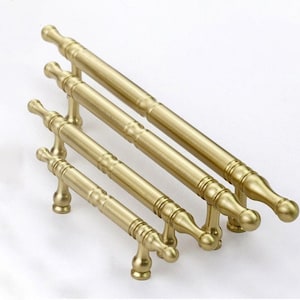 Könnte beinhalten: Vier goldfarbene Schrankgriffe mit einem dekorativen Design und abgerundeten Enden. Die Griffe sind versetzt angeordnet, wobei der größte Griff oben und der kleinste Griff unten ist.