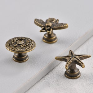 May include: Three antique bronze cabinet knobs with different designs. One knob features a circular design with the text "GREAT IND. & NOTHING", another knob features a bee design, and the third knob features a starfish design.