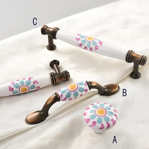 以下が含まれることがあります： ピンク、ブルー、イエローのフラワーデザインが施されたセラミック製のキャビネットノブ3個。ノブはブロンズ仕上げで、A、B、Cとラベル付けされています。
