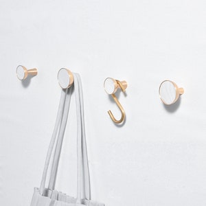 Könnte beinhalten: Drei goldene Haken mit weißen Marmorakzenten, die an einer weißen Wand montiert sind. Ein Haken hat eine gebogene Form, während die anderen beiden gerade sind. Eine weiße Einkaufstasche hängt an einem der geraden Haken.