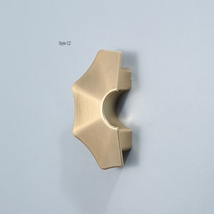 Peut inclure: Un bouton de porte de placard de forme géométrique, de couleur dorée, légèrement arrondi et incurvé. Le bouton est étiqueté "Style C2".