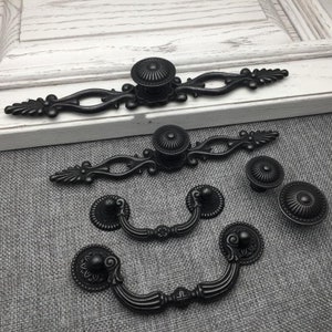 Könnte beinhalten: Ein Satz von sechs schwarzen Metall-Schrankknöpfen und -griffen mit verzierten Designs. Die Knöpfe sind rund mit einer erhabenen Mitte, und die Griffe sind in Form einer Schlaufe mit einem dekorativen Ende.