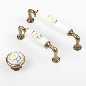 May include: Set of three ceramic cabinet knobs with a floral design. The knobs are white with a yellow flower and green leaves. The base of the knobs is a brass finish.