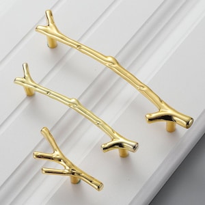 Op de afbeelding: Drie gouden kastknoppen in de vorm van takken. De knoppen zijn in verschillende maten en hebben een gladde, gepolijste afwerking.