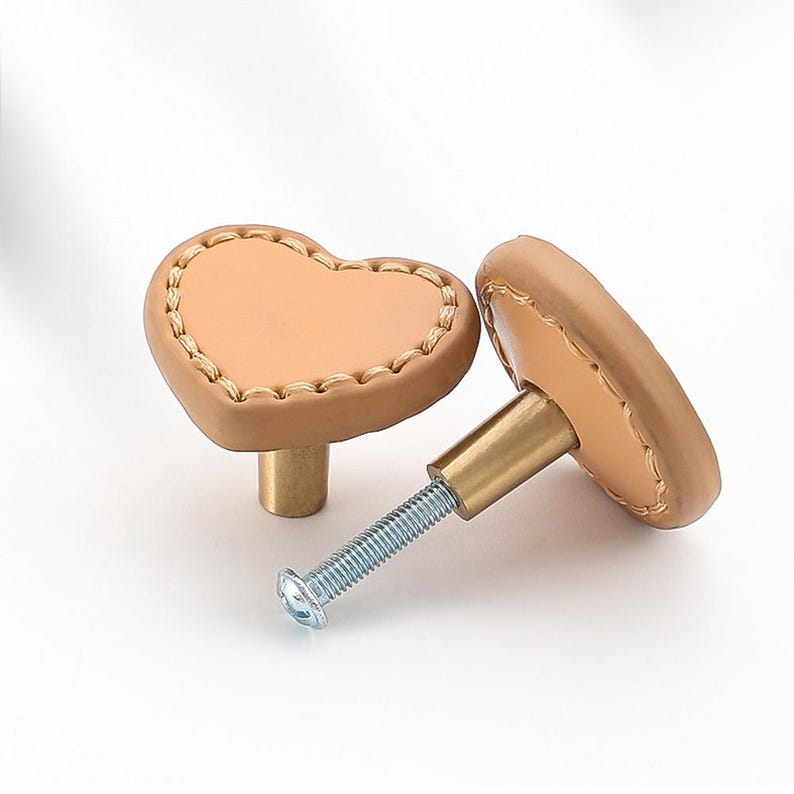 May include: Two heart-shaped cabinet knobs with a tan leather finish and gold-coloured metal hardware. The knobs have a stitched edge.