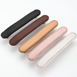 May include: Six leather drawer pulls in various colours, including black, brown, beige, pink, and white. Each pull has a stitched edge and a gold metal screw.