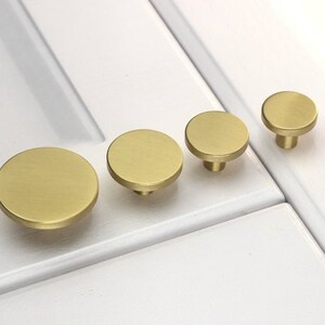 May include: Four round, brushed brass cabinet knobs of varying sizes. The knobs are shown on a white surface.