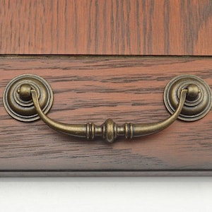 May include: Antique brass cabinet pull with a rounded, decorative design. The pull has a curved handle and two round knobs on either end.
