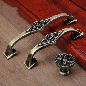 Könnte beinhalten: Zwei antike goldfarbene Schrankgriffe mit einem rautenförmigen Design und filigranen Verzierungen. Ein passender antiker goldfarbener Knopf mit einem ähnlichen Verzierungsdesign ist ebenfalls enthalten.