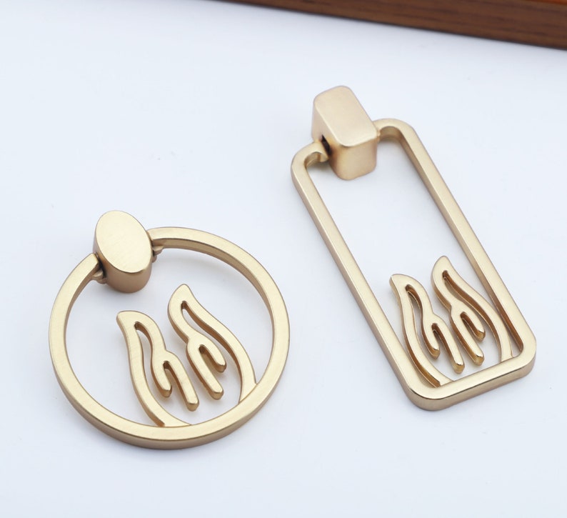 May include: Two gold-toned cabinet pulls with a flame design. One pull is a ring with a round knob, the other is a rectangular pull with a rounded top.