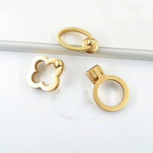 May include: Three gold cabinet knobs in different shapes: a round knob with a small handle, a four-leaf clover knob, and an oval knob with a small handle.