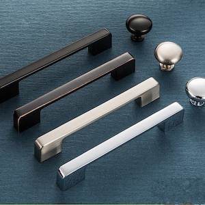 Può includere: Quattro maniglie per armadi in diverse finiture: nero, bronzo, nichel e cromo. Ogni maniglia ha una forma rettangolare con bordi arrotondati. Le maniglie sono disposte in fila da sinistra a destra, con la maniglia nera in alto e la maniglia cromata in basso.