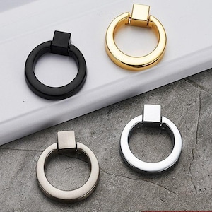 Könnte beinhalten: Vier verschiedene Schranktürgriffe in verschiedenen Ausführungen. Die Griffe sind alle rund mit einem quadratischen Knopf oben. Die Ausführungen umfassen Schwarz, Gold, Silber und Nickel.