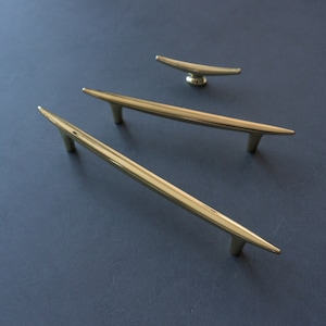 May include: Three gold-toned metal cabinet pulls with a modern, minimalist design. The pulls are in the shape of a thin, elongated triangle with a pointed tip.