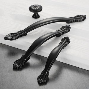 May include: A collection of black cabinet handles and a knob. The set includes two curved handles, a smaller handle, and a round knob. The handles feature decorative detailing. The hardware is presented on a white and grey surface.