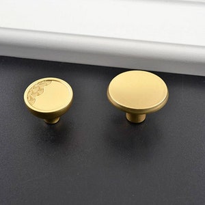 Puede incluir: Dos tiradores de armario de latón. Uno tiene un diseño redondo simple, mientras que el otro presenta un patrón decorativo. Los tiradores están sobre una superficie oscura, con una moldura blanca en el fondo. Ideales para muebles.